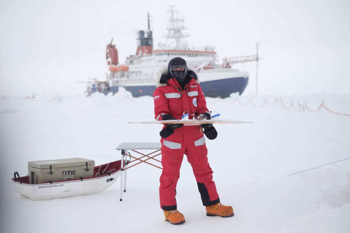 Gina Jozef holds a small uncrewed aerial system in her hand while standing on snow and ice. Directly behind her are a small table and boat with a hard cooler inside it. The R/V Polarstern is visible in the distance.