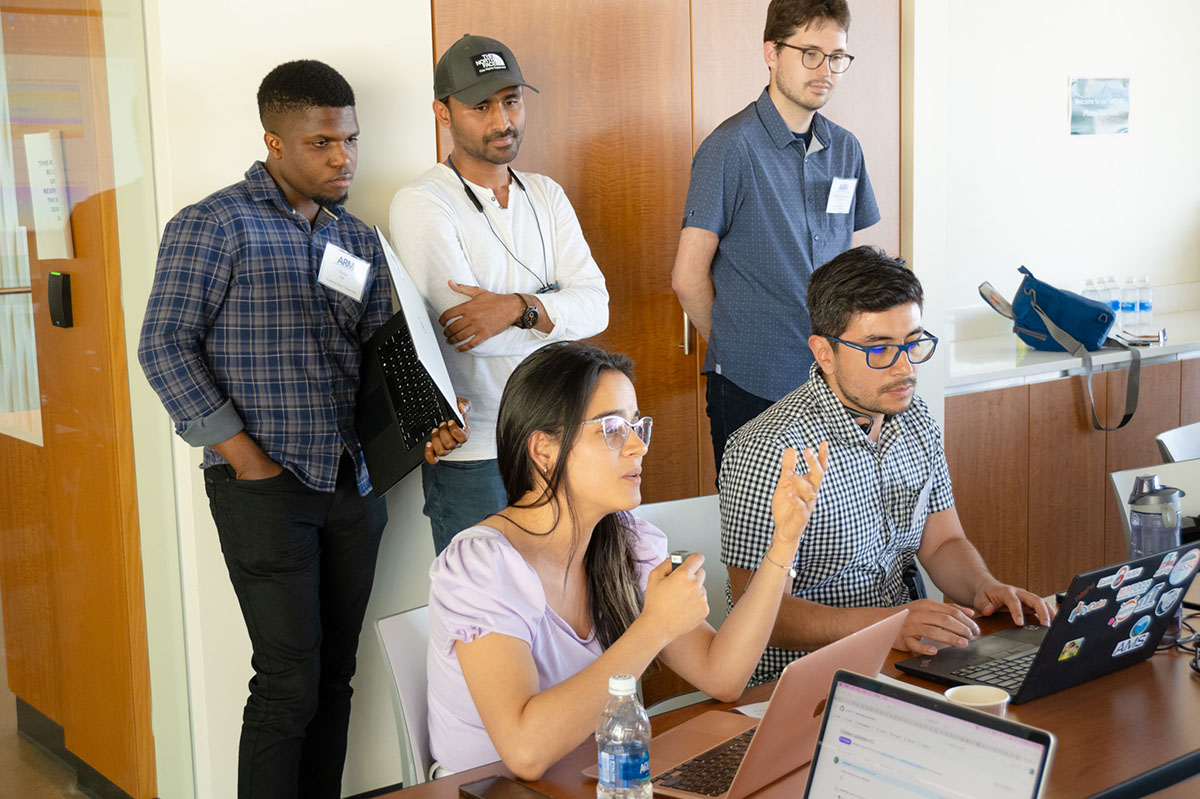 Two people are sitting down looking at laptops while their three teammates look on behind them.